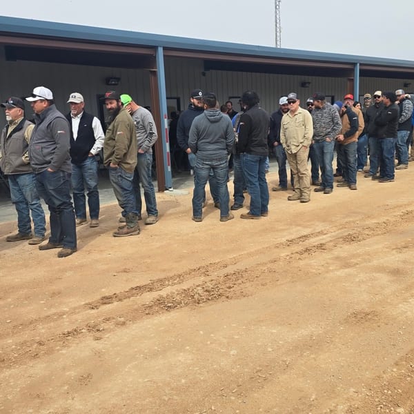 A large group of men in work clothes and hats stand in a line on a dirt road.