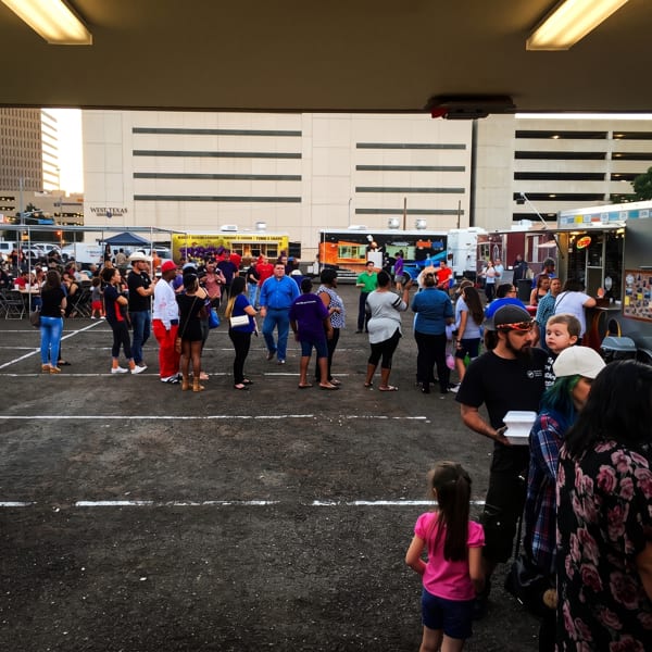 Crowd of people gathered around food trucks in a parking lot with city buildings in the background.