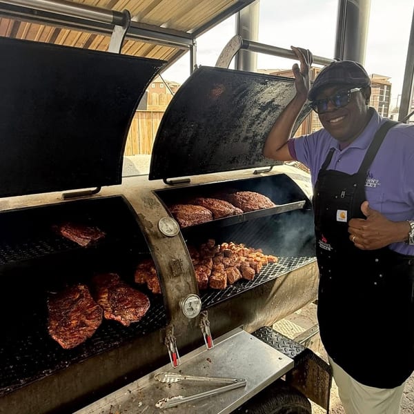 A smiling man in a purple shirt and apron stands next to a large smoker filled with barbecue.