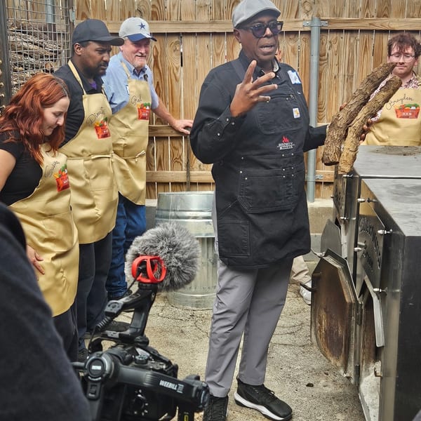 A man in an apron and hat gestures while holding wood, teaching a group outdoors.