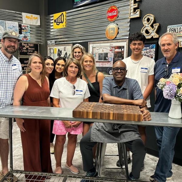 A group of ten people, including a chef, smiling behind a metal counter with flowers and a cutting board.